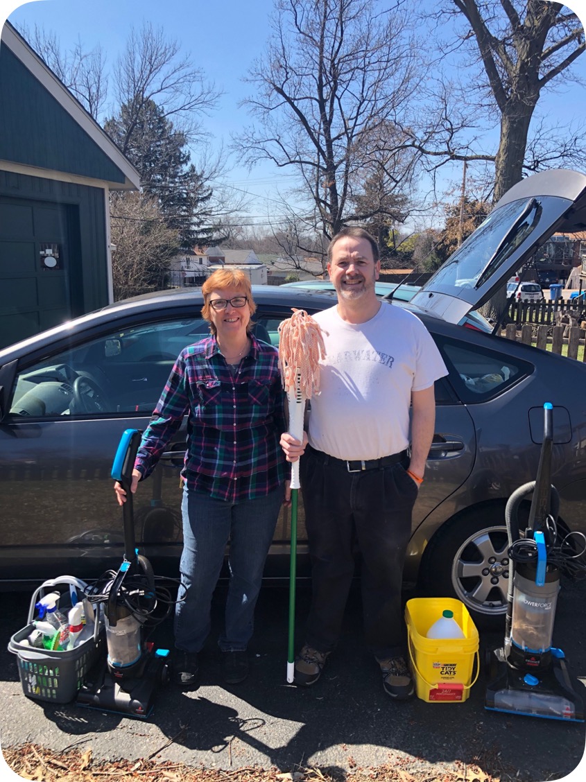 Two people posing with cleaning supplies in front of a parked vehicle and driveway